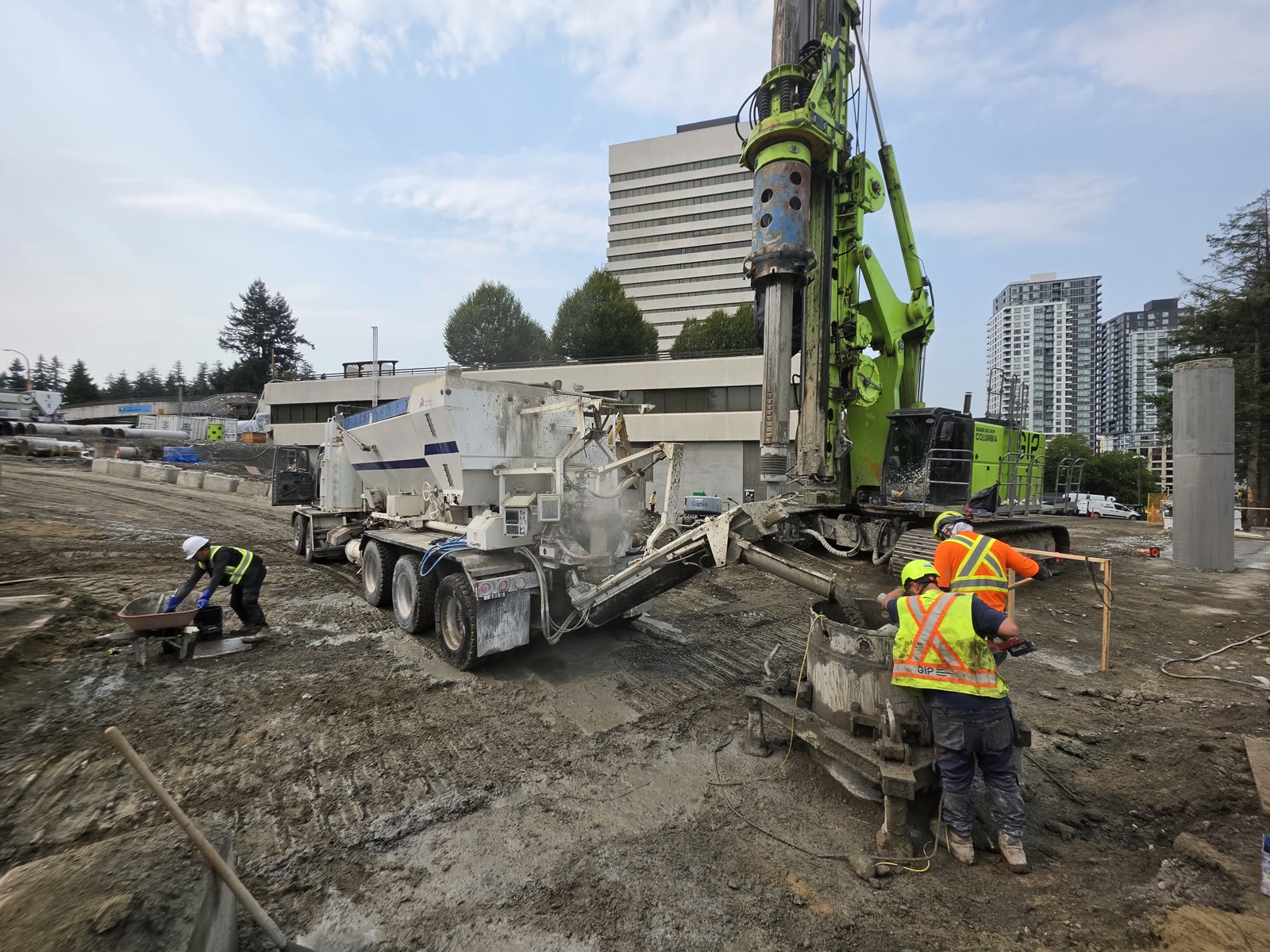 Crew placing concrete from a volumetric mixer beside a drilling rig on a city site