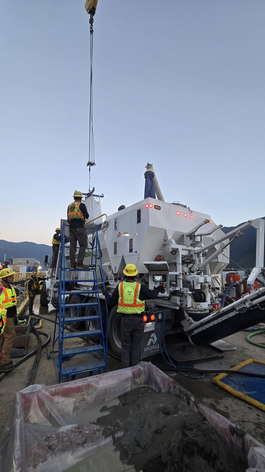 Sustainable Concrete crew running the volumetric mixer at dusk on the Woodfibre LNG site, with the crane line overhead and grout being placed